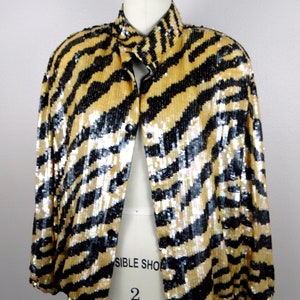 K&ouml;nnte beinhalten: Eine paillettenbesetzte Jacke in Gold und Schwarz mit einem auff&auml;lligen Zebrastreifenmuster. Die Jacke hat einen Kragen und wird mit Kn&ouml;pfen geschlossen. Das Kleidungsst&uuml;ck wird auf einer Schaufensterpuppe pr&auml;sentiert, um sein Design und seinen Stil hervorzuheben.