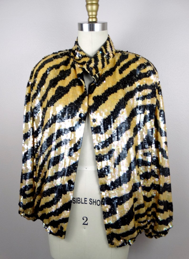K&ouml;nnte beinhalten: Eine paillettenbesetzte Jacke in Gold und Schwarz mit einem auff&auml;lligen Zebrastreifenmuster. Die Jacke hat einen Kragen und wird mit Kn&ouml;pfen geschlossen. Das Kleidungsst&uuml;ck wird auf einer Schaufensterpuppe pr&auml;sentiert, um sein Design und seinen Stil hervorzuheben.