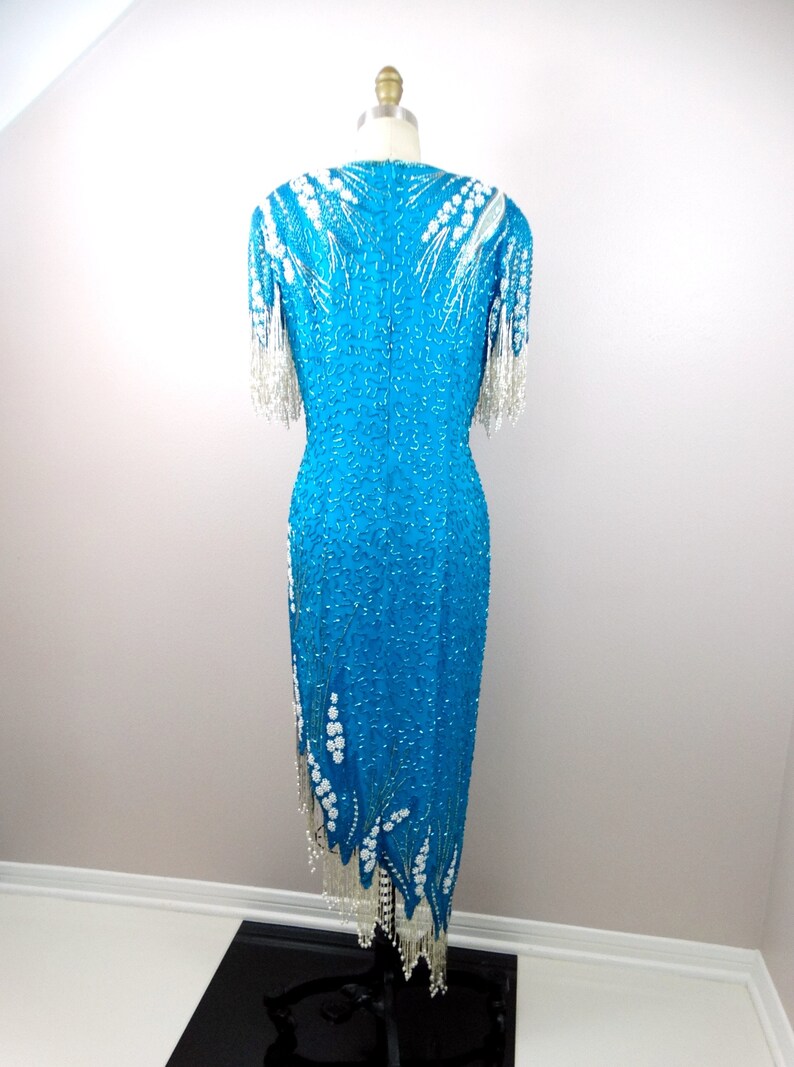 May include: A turquoise sequined dress with white beaded embellishments and fringe. The dress has a fitted bodice and a flowing skirt.