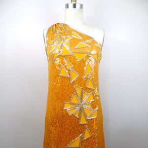 K&ouml;nnte beinhalten: Ein einseitiges Paillettenkleid in leuchtend gelber Farbe mit einem geometrischen Muster aus silbernen und gelben Dreiecken.