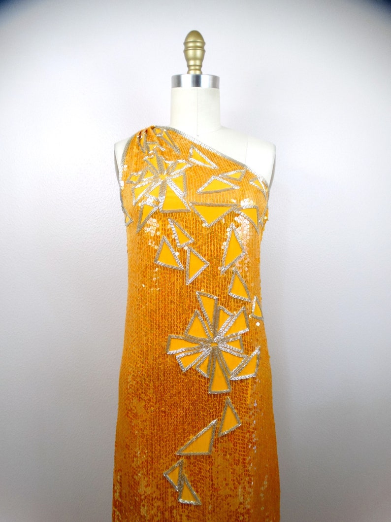 K&ouml;nnte beinhalten: Ein einseitiges Paillettenkleid in leuchtend gelber Farbe mit einem geometrischen Muster aus silbernen und gelben Dreiecken.