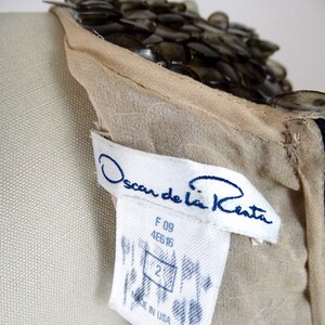 May include: A beige sheer dress with a beaded neckline. The dress has a white tag with the text "Oscar de la Renta" in blue lettering. The tag also includes the size "2" and the text "Made in USA".