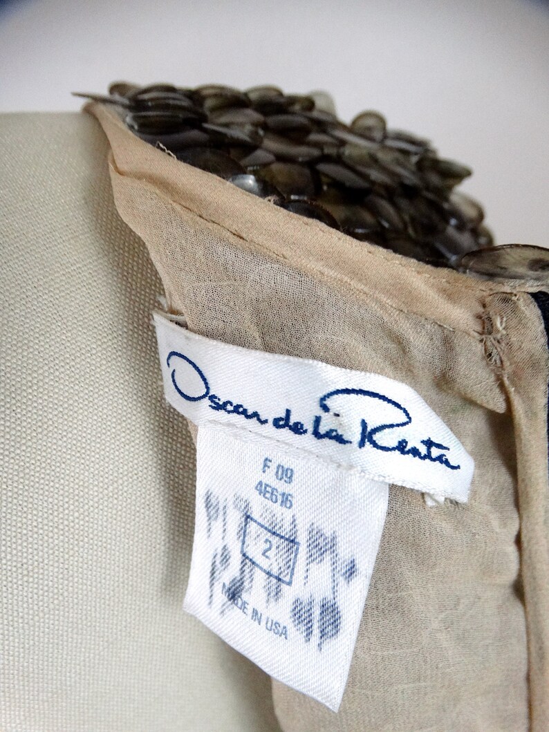 May include: A beige sheer dress with a beaded neckline. The dress has a white tag with the text "Oscar de la Renta" in blue lettering. The tag also includes the size "2" and the text "Made in USA".