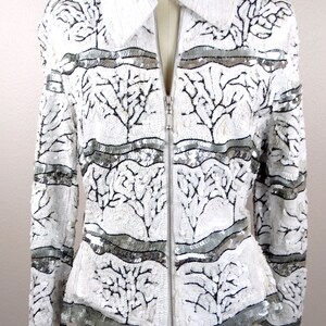 May include: White sequined jacket with a stand-up collar and a silver zipper. The jacket features a pattern of black tree-like designs and horizontal silver bands. A stylish piece of outerwear.