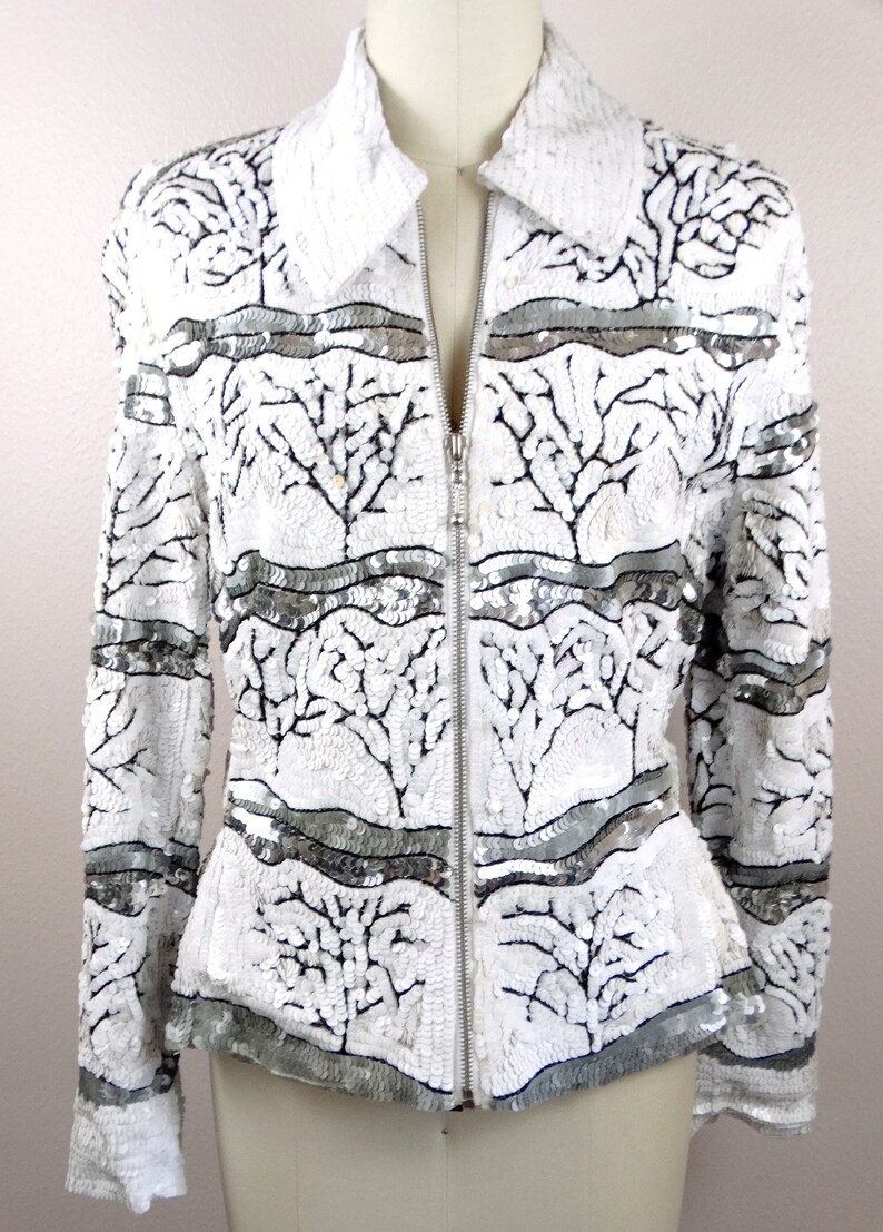 May include: White sequined jacket with a stand-up collar and a silver zipper. The jacket features a pattern of black tree-like designs and horizontal silver bands. A stylish piece of outerwear.