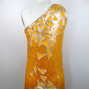 K&ouml;nnte beinhalten: Ein einseitiges Paillettenkleid in leuchtend gelber Farbe mit einem geometrischen Muster aus silbernen Pailletten. Das Kleid hat eine figurbetonte Silhouette und eine lange L&auml;nge.