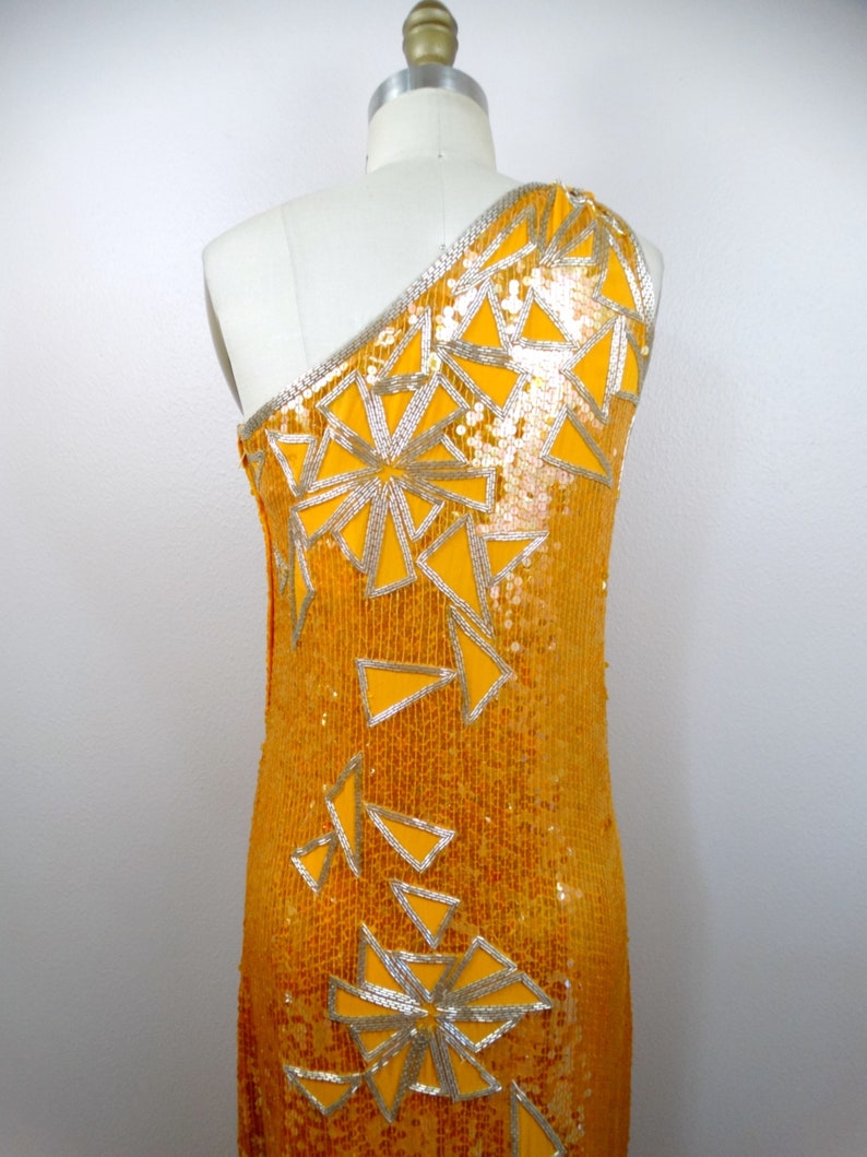 K&ouml;nnte beinhalten: Ein einseitiges Paillettenkleid in leuchtend gelber Farbe mit einem geometrischen Muster aus silbernen Pailletten. Das Kleid hat eine figurbetonte Silhouette und eine lange L&auml;nge.
