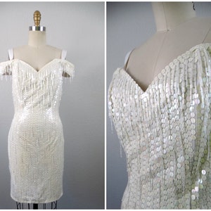 May include: A white off-the-shoulder dress with a sequined bodice and fringe detail. The dress is fitted and has a short length.