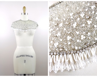 May include: A white beaded and sequined collar with a fringe trim. The collar is displayed on a white dress form.