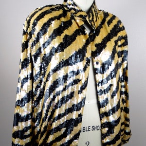 K&ouml;nnte beinhalten: Gold-schwarze Paillettenjacke mit Streifenmuster. Die Jacke wird auf einer Schaufensterpuppe pr&auml;sentiert und hat einen Kragen und schwarze Kn&ouml;pfe. Die Pailletten erzeugen einen schimmernden Effekt und verleihen dem Kleidungsst&uuml;ck einen Hauch von Glamour.