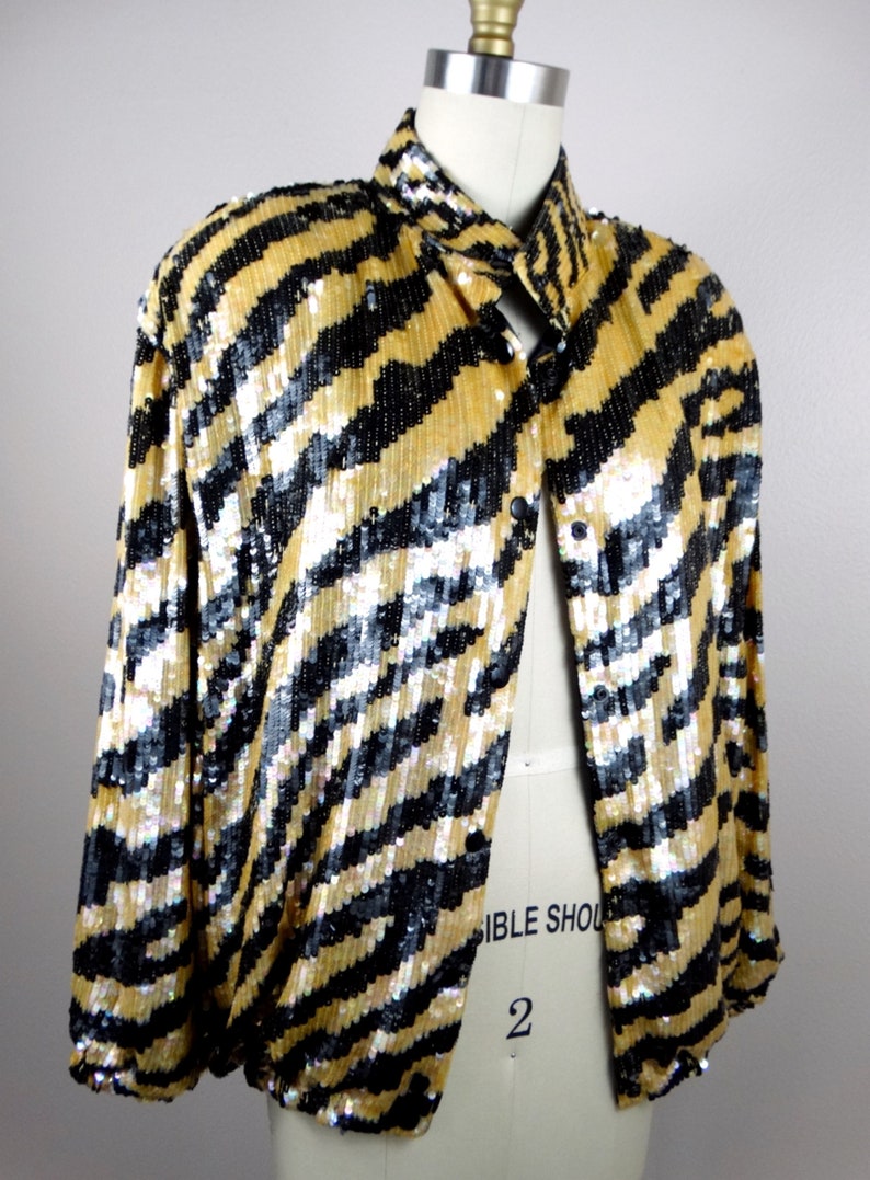 K&ouml;nnte beinhalten: Gold-schwarze Paillettenjacke mit Streifenmuster. Die Jacke wird auf einer Schaufensterpuppe pr&auml;sentiert und hat einen Kragen und schwarze Kn&ouml;pfe. Die Pailletten erzeugen einen schimmernden Effekt und verleihen dem Kleidungsst&uuml;ck einen Hauch von Glamour.