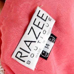 Op de afbeelding: Een wit label met zwarte tekst dat "Riazee Boutique Made in India" zegt met een zwart label eronder dat "M.F. 14" zegt en een maat 14.