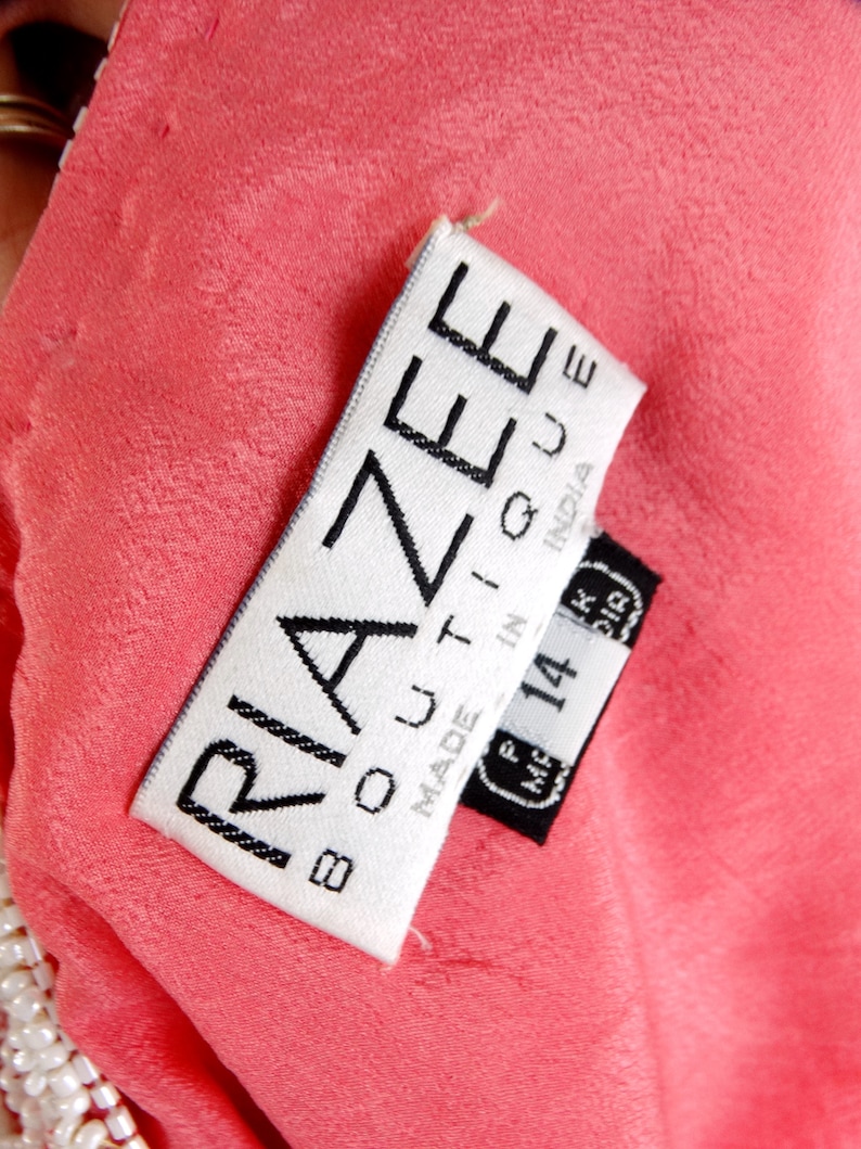 Op de afbeelding: Een wit label met zwarte tekst dat "Riazee Boutique Made in India" zegt met een zwart label eronder dat "M.F. 14" zegt en een maat 14.