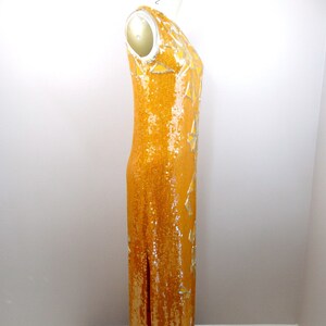 K&ouml;nnte beinhalten: Ein langes, &auml;rmelloses, paillettenbesetztes Kleid in leuchtend gelber Farbe mit silbernen Akzenten. Das Kleid hat einen Schlitz an der Seite und eine figurbetonte Silhouette.