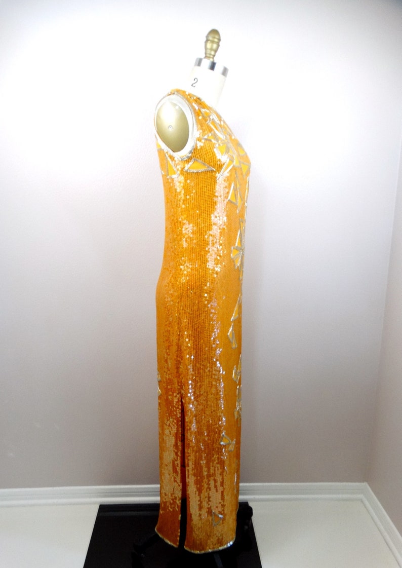 K&ouml;nnte beinhalten: Ein langes, &auml;rmelloses, paillettenbesetztes Kleid in leuchtend gelber Farbe mit silbernen Akzenten. Das Kleid hat einen Schlitz an der Seite und eine figurbetonte Silhouette.