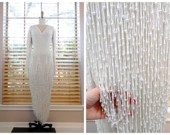 Puede incluir: Un vestido blanco de cuentas con mangas largas y escote en V. El vestido está hecho de una tela fina y delicada y está cubierto de pequeñas cuentas blancas. Las cuentas están dispuestas en un patrón que crea un efecto de flecos.