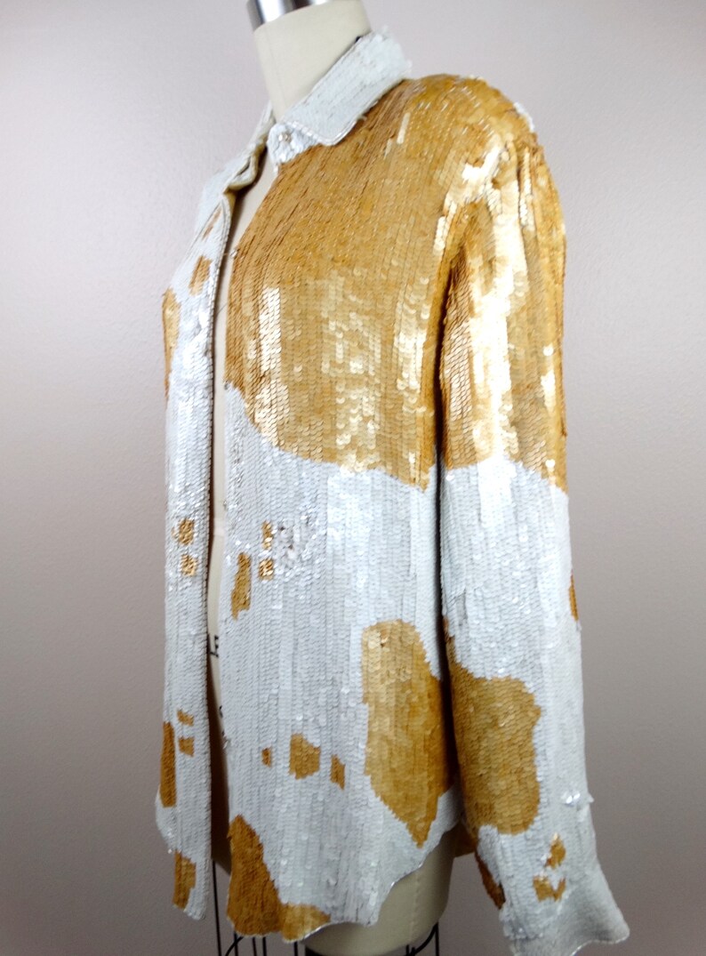 May include: A white and gold sequined button-up shirt with a long sleeve. The shirt has a geometric pattern with large white and gold shapes.