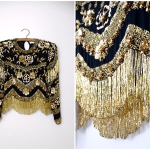 Puede incluir: Una blusa negra y dorada con lentejuelas, mangas largas y ribete de flecos. La blusa está decorada con bordados florales y cuentas.