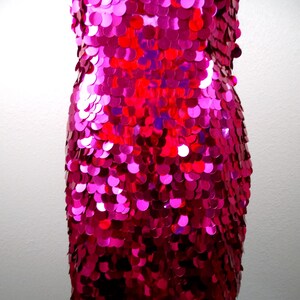 May include: A sleeveless, knee-length dress covered in vibrant, fuchsia-colored sequins. The sequins are round and densely packed, creating a shimmering effect. The dress is displayed on a black dress form.