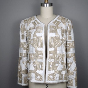 May include: A white and beige beaded jacket with a geometric pattern. The jacket has a long sleeve and a open front.