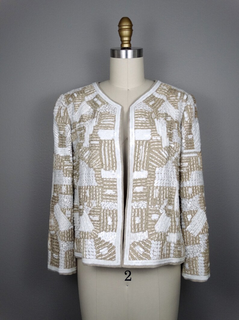 May include: A white and beige beaded jacket with a geometric pattern. The jacket has a long sleeve and a open front.