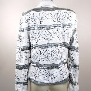 May include: White sequined jacket with a gray collar and cuffs. The jacket features a pattern of black tree-like designs and horizontal gray bands. The garment is displayed on a mannequin, showcasing its back.