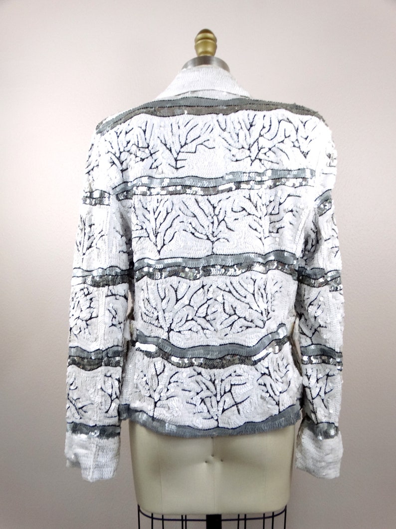 May include: White sequined jacket with a gray collar and cuffs. The jacket features a pattern of black tree-like designs and horizontal gray bands. The garment is displayed on a mannequin, showcasing its back.