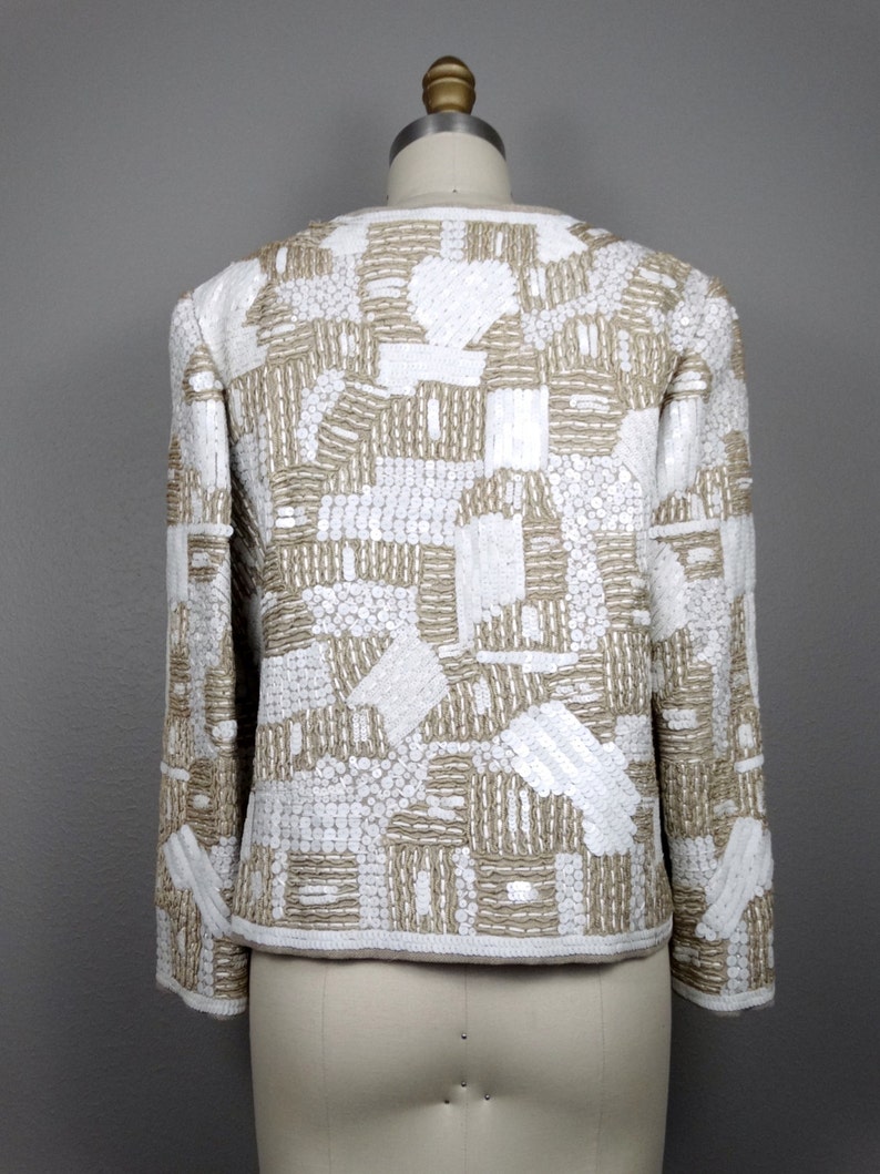 May include: A white and gold sequined jacket with a geometric pattern. The jacket has long sleeves and a cropped length.