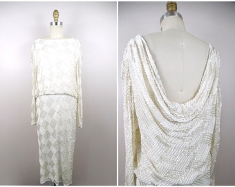 May include: A white beaded dress with long sleeves and a draped back. The dress has a geometric pattern and is made of a soft, flowing fabric.