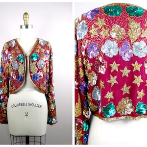May include: A cropped sequined jacket with a red base and colorful floral and star embellishments. The jacket has a V-neck and long sleeves. The label reads "Collapsible Shoulder 2".