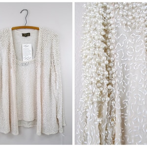 Puede incluir: Un cárdigan blanco de manga larga y una blusa a juego con cuello redondo. El cárdigan está adornado con cuentas de imitación de perlas.
