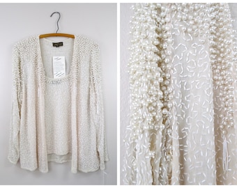 Puede incluir: Un cárdigan blanco de manga larga y una blusa a juego con cuello redondo. El cárdigan está adornado con cuentas de imitación de perlas.