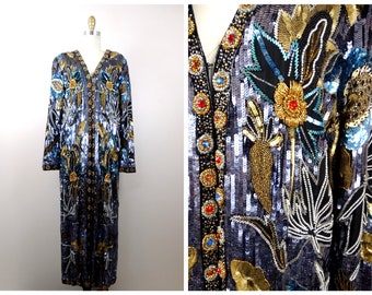 Op de afbeelding: Een lange, met pailletten bezette robe met een V-hals en bloemenborduurwerk. De robe is zwart met zilveren, gouden en blauwe pailletten. Het borduurwerk toont bloemen, bladeren en andere botanische motieven.