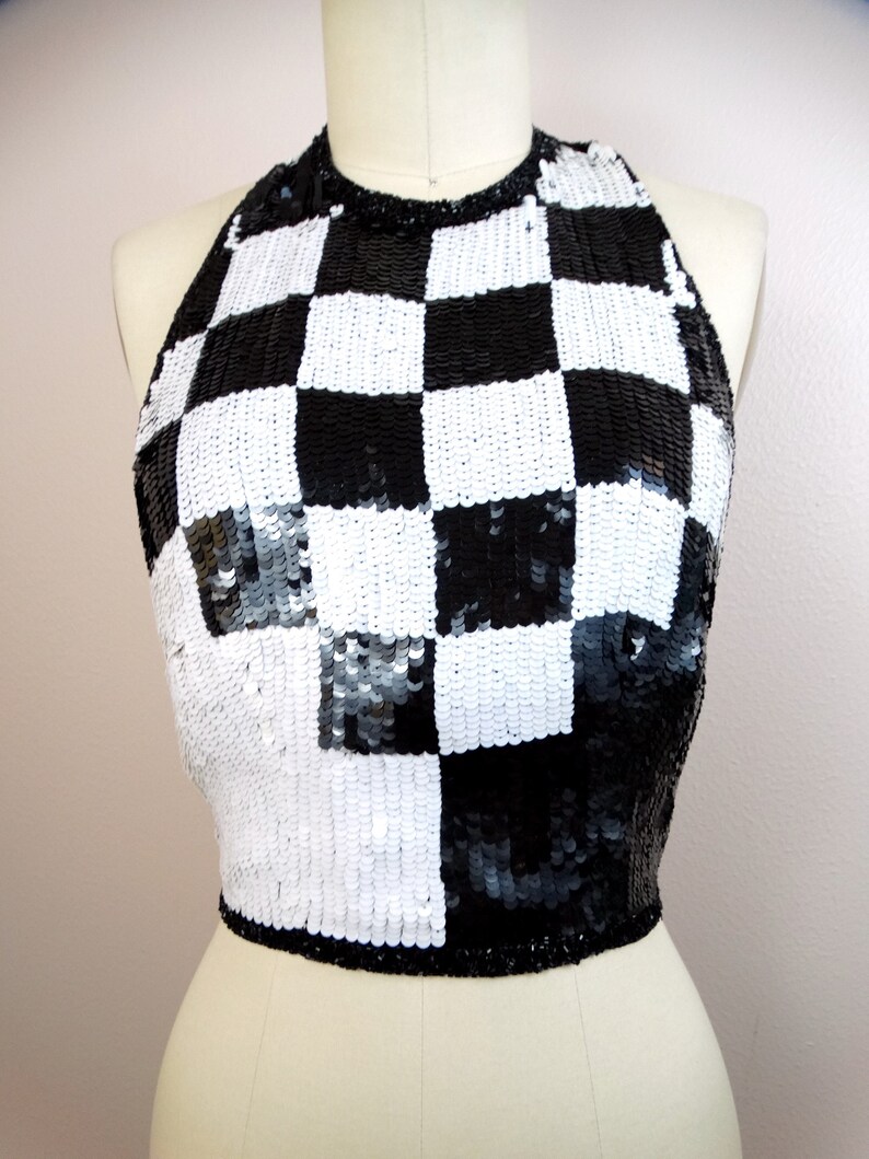 May include: A black and white checkered halter top made of sequins. The top is cropped and has a halter neckline.