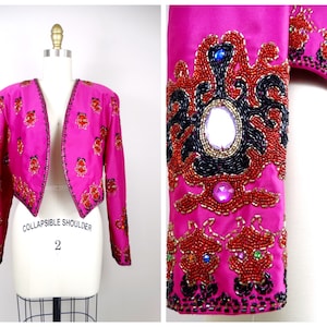 Puede incluir: Una chaqueta rosa corta con un detalle de hombro plegable. La chaqueta está adornada con cuentas y lentejuelas rojas, negras y doradas. La chaqueta tiene cuello en V y mangas largas.