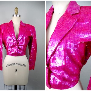 May include: A hot pink sequined blazer with a classic notched lapel and a single-button closure. The cropped jacket is displayed on a mannequin, showcasing its tailored fit and sparkling texture. The garment is a statement piece.
