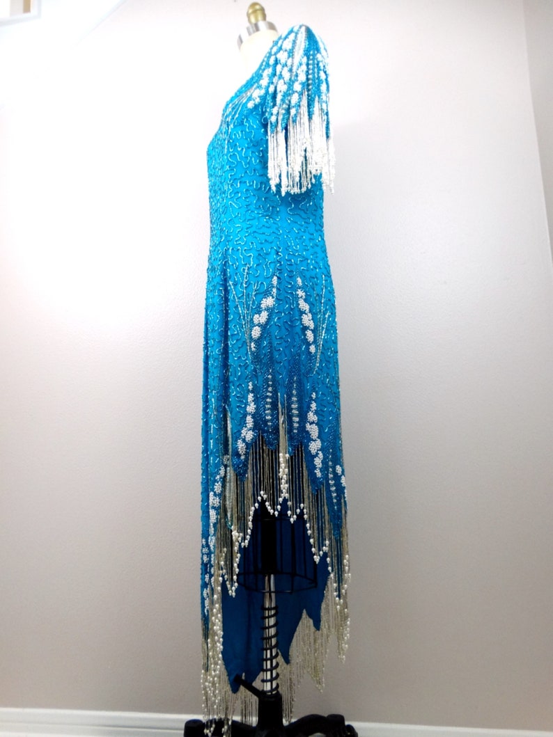 May include: A turquoise sequined dress with white beaded fringe. The dress has a scoop neck and short sleeves. The fringe is on the sleeves, hem, and neckline.