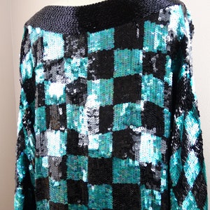 Puede incluir: Una blusa negra y turquesa con lentejuelas y un patr&oacute;n de cuadros. La blusa tiene cuello de barco y mangas largas.