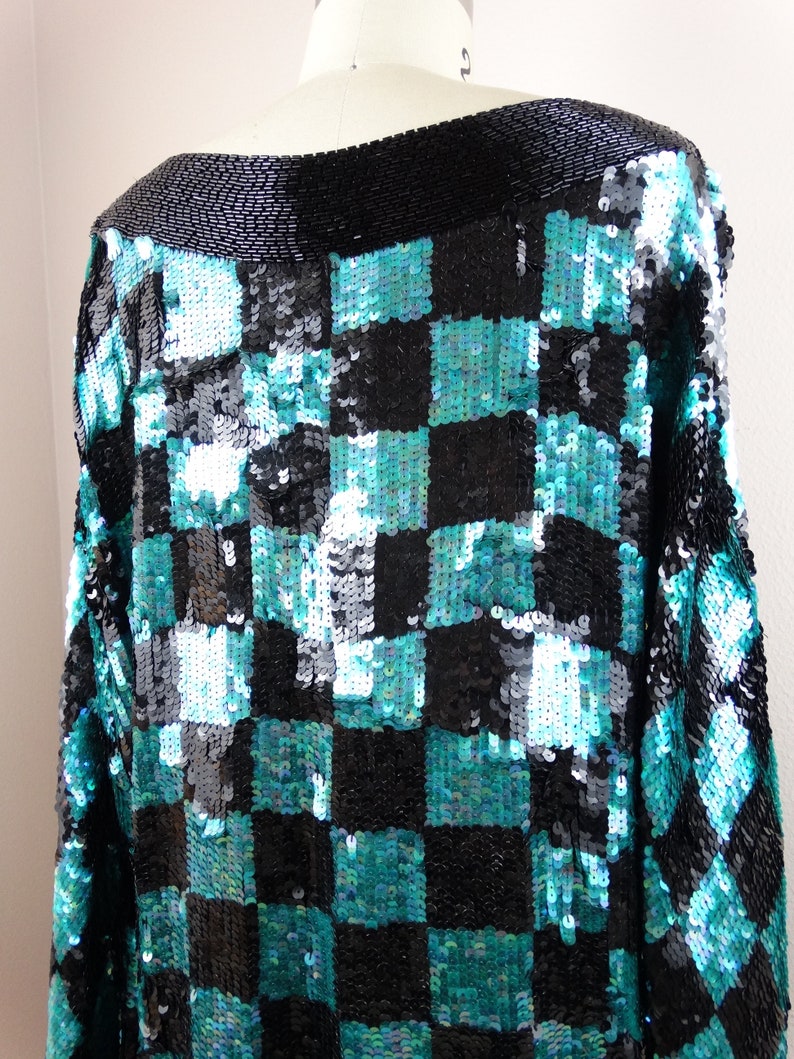 Puede incluir: Una blusa negra y turquesa con lentejuelas y un patr&oacute;n de cuadros. La blusa tiene cuello de barco y mangas largas.