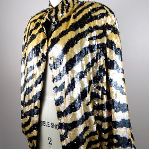 K&ouml;nnte beinhalten: Eine Paillettenjacke mit einem schwarz-goldenen Zebrastreifenmuster. Die Jacke hat einen Stehkragen und lange &Auml;rmel. Das Kleidungsst&uuml;ck wird auf einer Schaufensterpuppe pr&auml;sentiert.