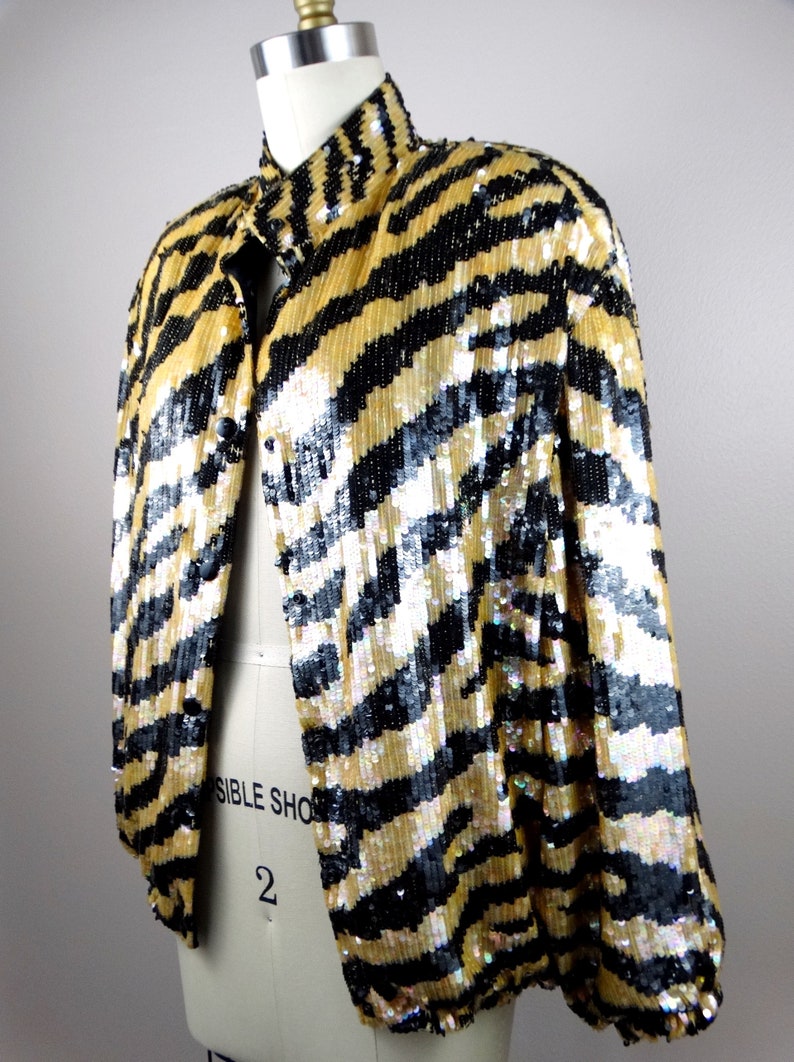 K&ouml;nnte beinhalten: Eine Paillettenjacke mit einem schwarz-goldenen Zebrastreifenmuster. Die Jacke hat einen Stehkragen und lange &Auml;rmel. Das Kleidungsst&uuml;ck wird auf einer Schaufensterpuppe pr&auml;sentiert.