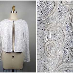 Puede incluir: Chaqueta blanca con frente abierto y mangas largas, exhibida en un maniquí. La chaqueta está muy adornada con cuentas y lentejuelas blancas, creando un diseño texturizado y ornamentado. El primer plano revela patrones intrincados.
