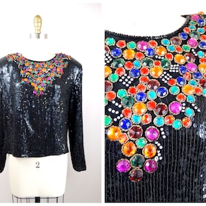May include: Black sequined long-sleeve top with a jewel-embellished neckline. The neckline features a colorful array of large, round gemstones in shades of red, orange, yellow, green, blue, and purple. The top is displayed on a mannequin.