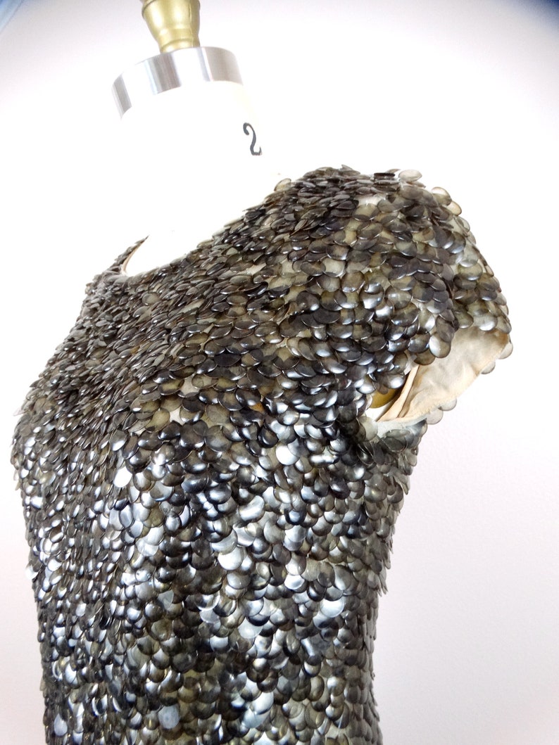 May include: A close-up of a silver and black dress with a unique design made of overlapping scales. The dress is on a white mannequin.