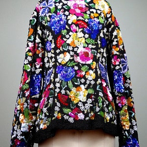 Pode incluir: Uma jaqueta preta de lantejoulas com um padr&atilde;o floral em tons de rosa, azul, verde, amarelo e vermelho. A jaqueta tem mangas compridas e um comprimento curto.