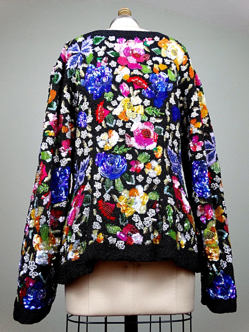 Pode incluir: Uma jaqueta preta de lantejoulas com um padr&atilde;o floral em tons de rosa, azul, verde, amarelo e vermelho. A jaqueta tem mangas compridas e um comprimento curto.
