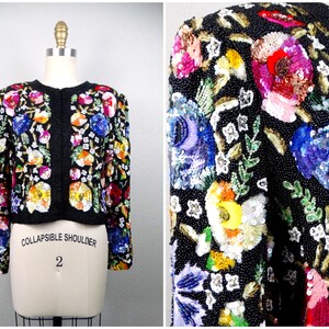 Peut inclure: Une veste noire en sequins avec un motif floral composé de fleurs, de feuilles et de sequins colorés. La veste a une fermeture à boutons et une étiquette 'Collapsible Shoulder'.
