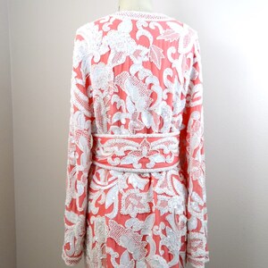 Op de afbeelding: Een lang, koraalrood jasje met een wit bloemenpatroon. Het jasje is versierd met witte pailletten en kralen. Het jasje heeft een riem in de taille.