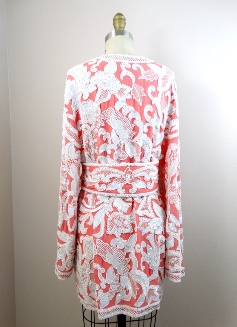 Op de afbeelding: Een lang, koraalrood jasje met een wit bloemenpatroon. Het jasje is versierd met witte pailletten en kralen. Het jasje heeft een riem in de taille.