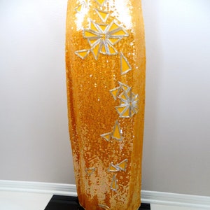 K&ouml;nnte beinhalten: Ein langes, goldenes Paillettenkleid mit einem silbernen geometrischen Muster. Das Kleid hat ein figurbetontes Oberteil und einen flie&szlig;enden Rock.